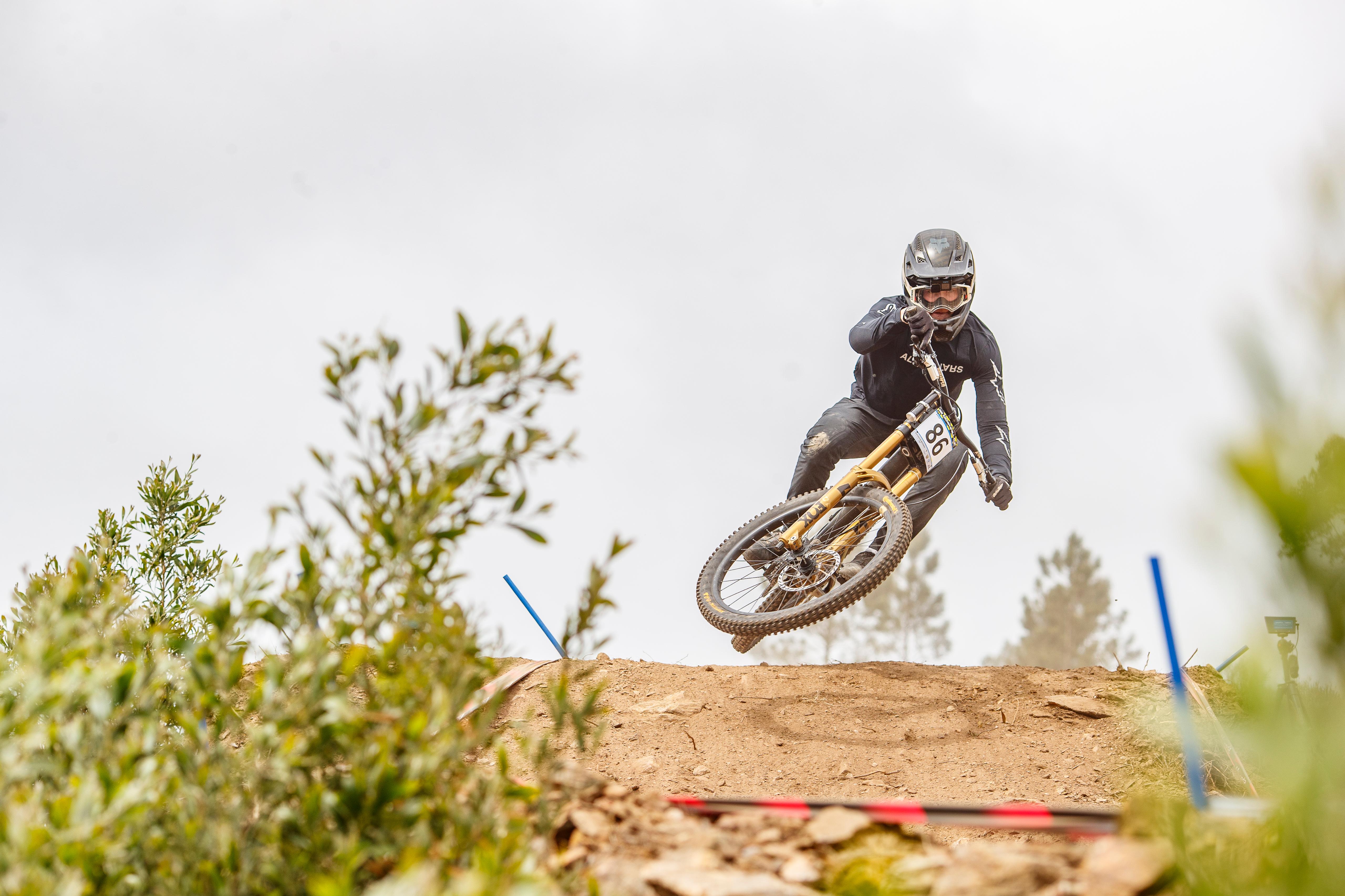 Louis Giller en plein saut lors d'une descente VTT (DH), dossard de course visible, terrain de terre et poussière.